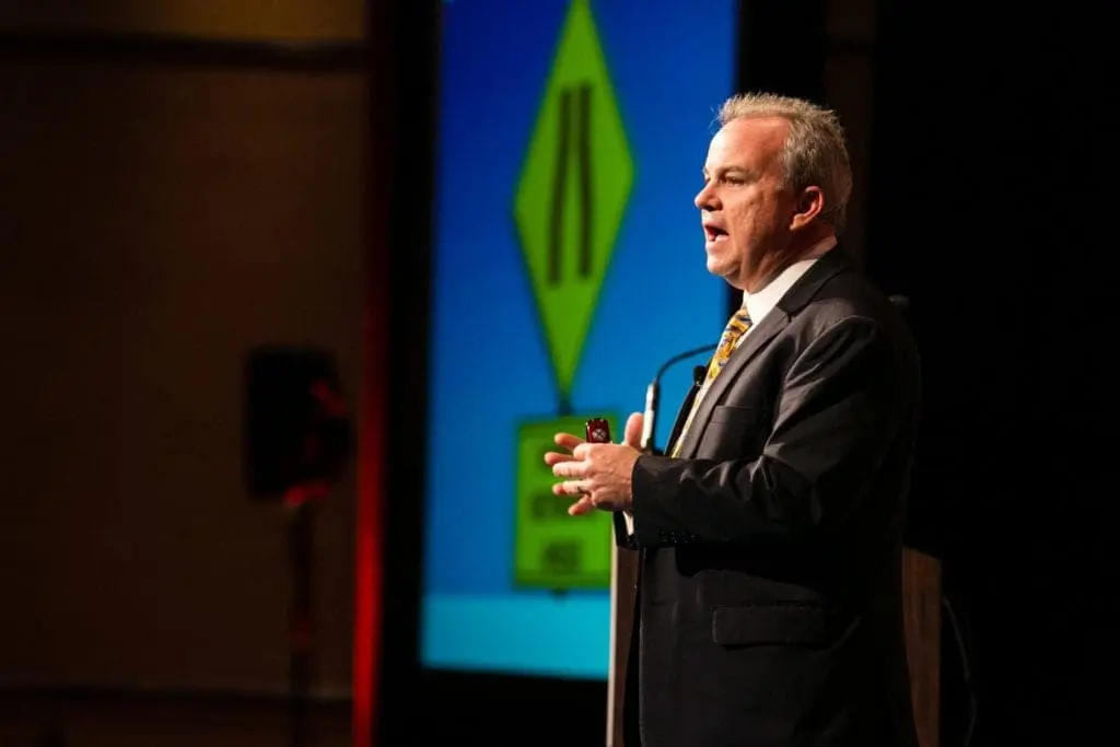 Corporate Event Photography1-14 A man in a suit speaks on stage at a corporate event, with a blue and green presentation slide in the background. The lighting focuses on him as he gestures with his hands, capturing the essence of professional headshots through skilled corporate event photography.