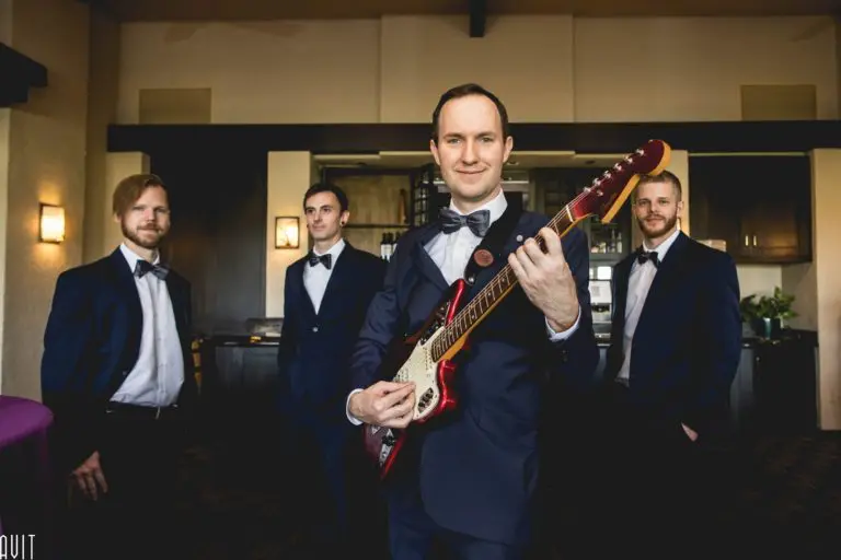 Four men in tuxedos stand indoors at a corporate event; the man in front smiles while holding an electric guitar. The three men behind him pose casually, all dressed formally in a warmly lit room.