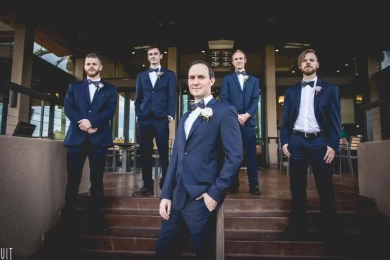 Five men in navy blue suits and bow ties pose confidently on outdoor steps, as if arriving for a seminar, with one standing in front and the others behind him outside a modern building with large windows.
