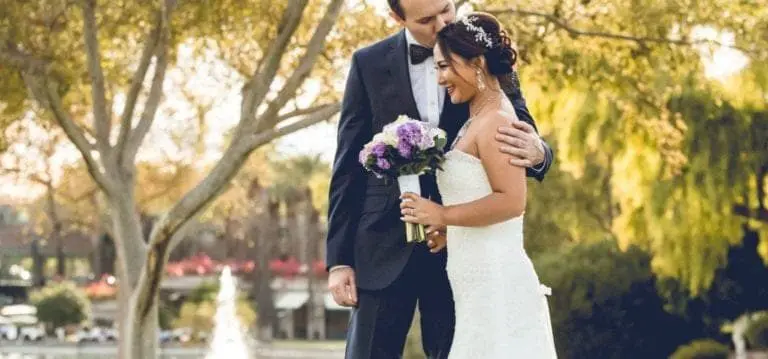 A groom in a dark suit gently kisses his smiling bride, who is holding a bouquet of purple flowers, outdoors in a sunlit park—an enchanting scene that could grace any seminar or corporate event celebrating love and new beginnings.