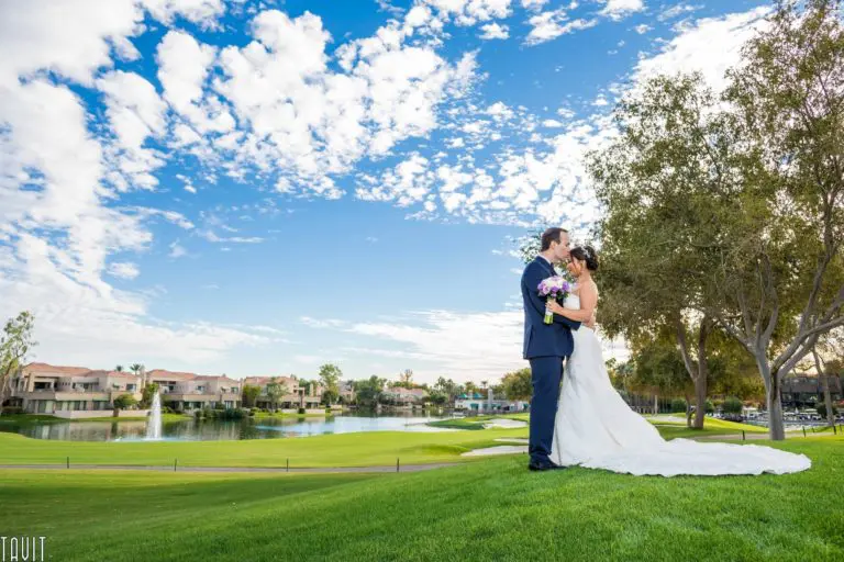 A bride and groom embrace on a lush green lawn near a pond, where houses and trees stand in the background—an enchanting spot that could host an intimate seminar or elegant corporate event under blue skies dotted with white clouds.