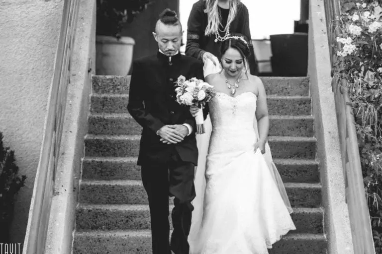 A bride in a strapless gown holding a bouquet walks down steps beside a groom in a dark suit. Guided by another person, they evoke the formality and joy of a special day—much like the poised entrance at a seminar or corporate event. Black and white photo.