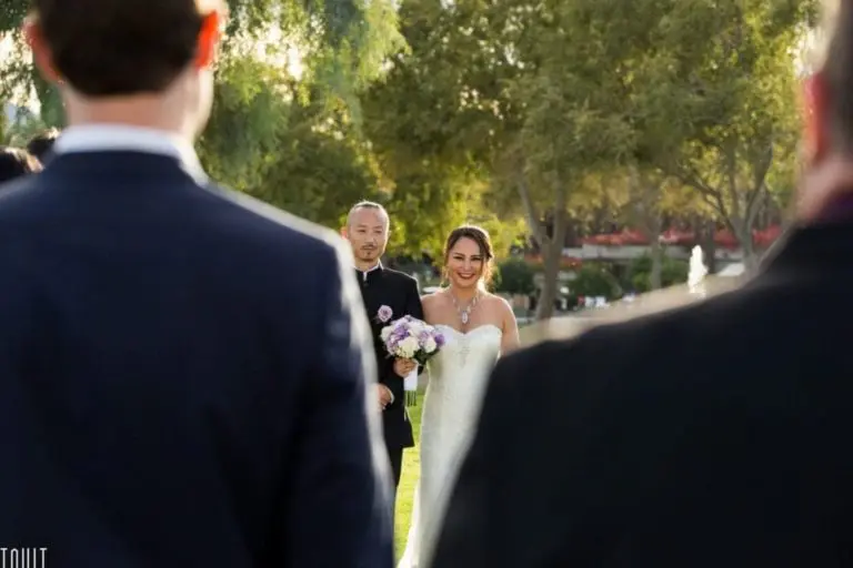 A smiling bride in a white dress holds a bouquet as she walks down the aisle outdoors with a man in a suit; guests in formal wear, reminiscent of a seminar or corporate event, are visible. Trees and greenery form the background.