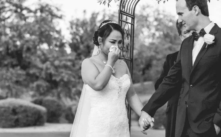A bride wipes tears from her face while holding hands with the groom during an outdoor ceremony; both are dressed formally, bringing heartfelt emotion to a setting usually reserved for a seminar or corporate event.