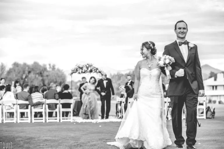 A bride in a strapless gown and a man in a suit walk arm-in-arm outdoors, smiling, with wedding guests seated and an arch visible in the background, creating a timeless scene far from any seminar or corporate event. The image is in black and white.