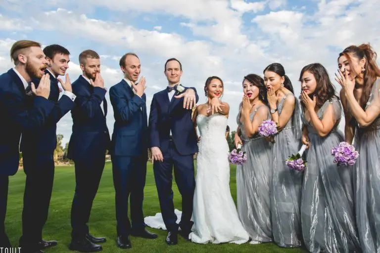 A bride and groom stand outdoors with their wedding party, playfully showing off their rings as if unveiling them at a corporate event. Bridesmaids in gray dresses and groomsmen in navy suits pose with exaggerated surprise under a partly cloudy sky.