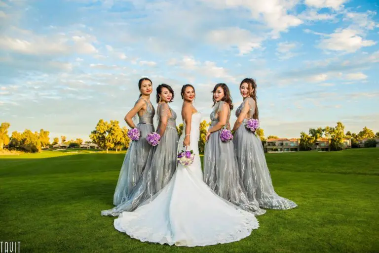 Five women pose outdoors on a green lawn under a blue sky with clouds. The bride wears a white gown and holds a bouquet, while four bridesmaids in gray dresses hold purple flowers, reminiscent of an elegant corporate event.