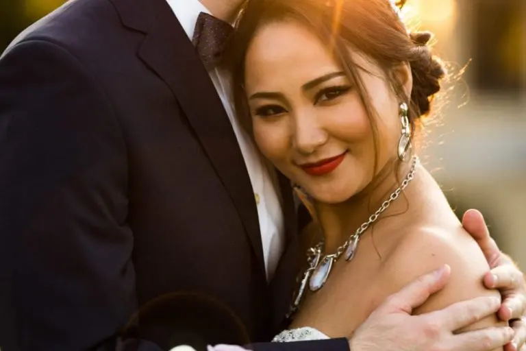 A bride and groom embrace outdoors at sunset, far from any seminar or corporate event. The smiling bride holds a bouquet of white and purple flowers as she leans into the groom, bathed in the warm glow of golden sunlight.