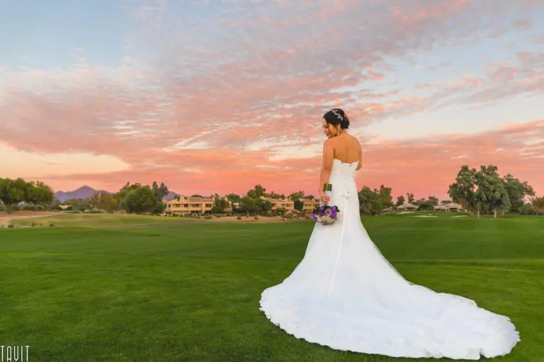 A bride in a white dress stands on a green lawn, holding a purple bouquet. She faces away under a colorful sunset sky, with houses and trees in the background, as if pausing before an intimate seminar or corporate event.