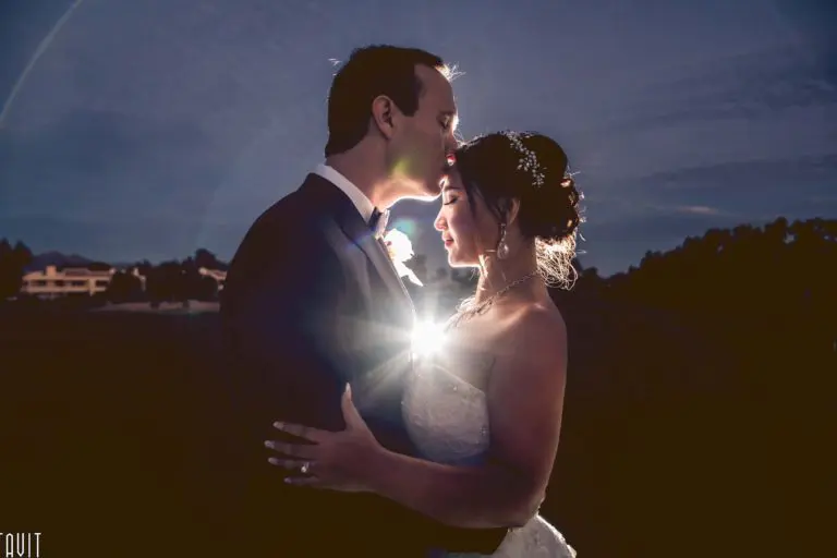 A groom in a tuxedo gently kisses the forehead of his bride in a white dress at dusk, while the soft, romantic glow between them feels worlds away from any seminar or corporate event.