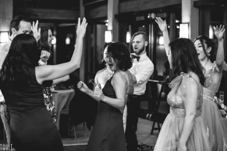 A group of people dressed formally are dancing enthusiastically, with their arms raised and smiles on their faces, at what appears to be a lively indoor corporate event or celebration. The photo is in black and white.