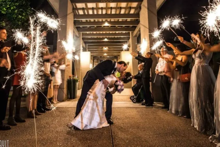A bride and groom share a kiss during an outdoor night celebration, where guests—fresh from a seminar or corporate event—hold sparklers and cheer as the couple walks down a path beneath a covered walkway.