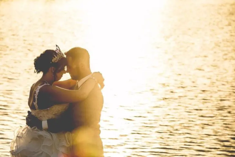A couple in wedding attire embraces closely by the water at sunset, the sun casting a golden glow and creating a romantic, backlit silhouette—capturing intimacy and elegance perfect for any seminar or corporate event inspiration.