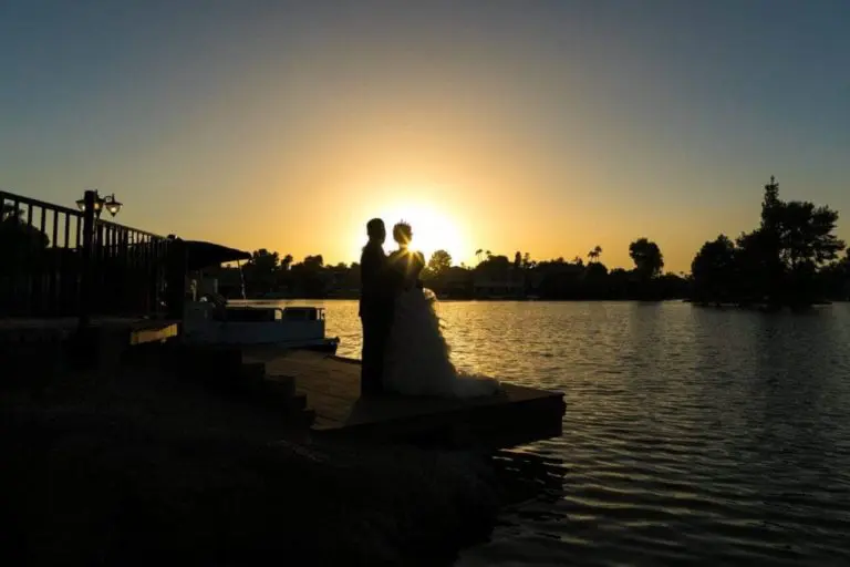 A bride and groom stand together on a dock at sunset, silhouetted against the golden sky, as if sharing a quiet moment away from the bustle of a seminar or corporate event, with a calm lake and trees in the background.