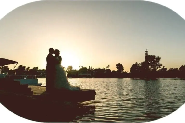 Silhouette of a bride and groom standing on a dock by a lake at sunset, embracing with the sun setting behind them, trees and houses in the background—a romantic scene perfect for weddings or even serene corporate event backdrops.