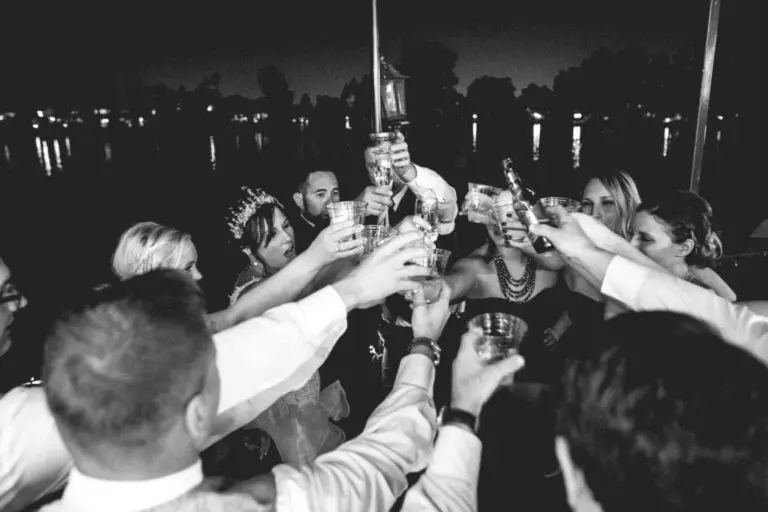 A group of people, including a bride in a tiara, raise their glasses together in a celebratory toast at an evening corporate event by the water. The image is in black and white.