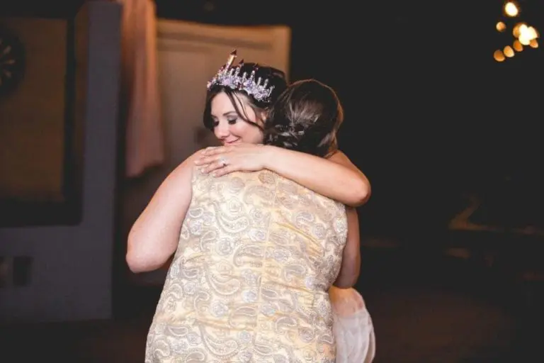 Two women are embracing warmly at a corporate event. One wears a sparkling crown and a patterned dress, smiling with her eyes closed. The scene is softly lit, creating a warm and intimate atmosphere.