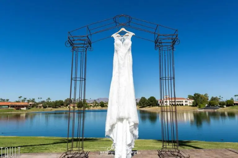 A white wedding dress hangs on a wrought iron archway against a backdrop of a serene lake, green lawns, and clear blue sky, with buildings and trees in the distance.