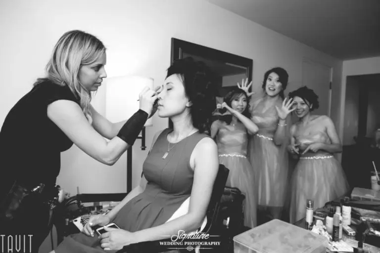 A woman is having her makeup done by a makeup artist, while four bridesmaids in matching dresses stand in the background, playfully posing and smiling. The room is filled with beauty products and the atmosphere is joyful.