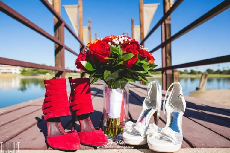 Red high heels and white sandals are placed next to a vase of red roses on a wooden bridge, with a serene lake in the background under a clear blue sky.
