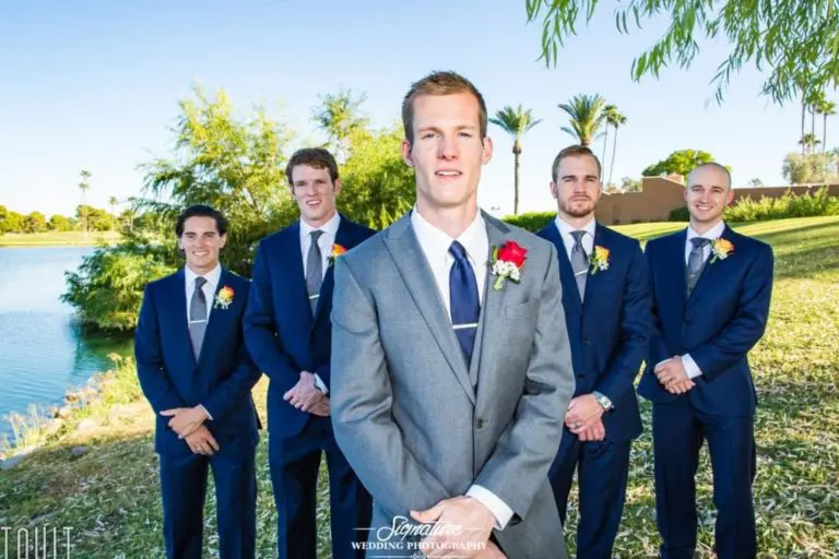 A groom in a gray suit stands in front of four groomsmen wearing dark blue suits and colorful boutonnieres. They are outdoors by a lake with grass and palm trees in the background. The sky is clear and sunny.