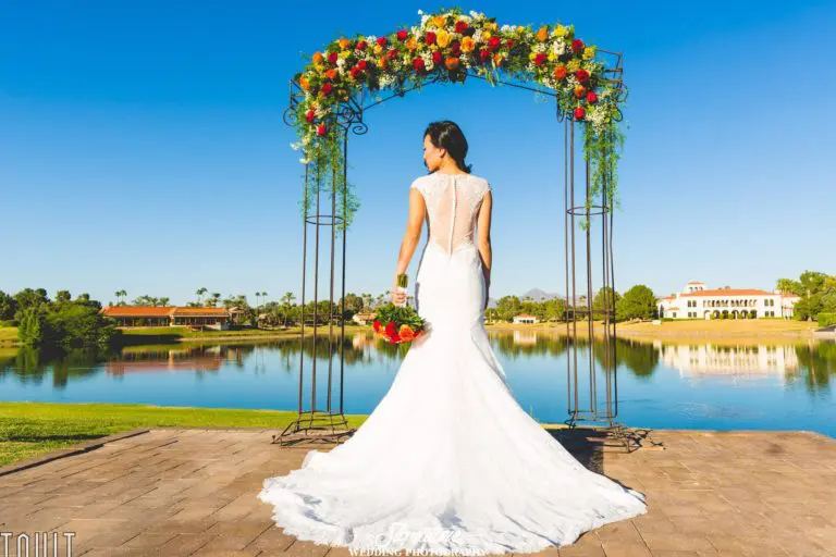 A bride in a white lace wedding gown stands on a paved path facing a lake. She's holding a bouquet of flowers and is framed by a floral arch. The background features a clear blue sky, greenery, and a distant building.