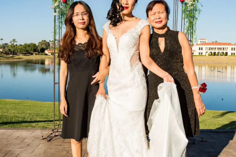 A bride in a white wedding dress stands between two women in black dresses. They pose in front of a flower-adorned arch with a lake and building in the background under a clear blue sky.