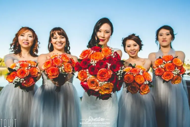 Five women in gray dresses hold bouquets of vibrant orange and red roses. They are smiling and standing outdoors against a clear blue sky. The central woman wears a white dress, indicating she is likely the bride.