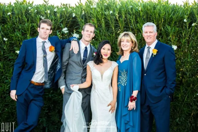 A smiling bride in a white dress poses with two men in suits, a woman in a teal dress, and another man in a blue suit, all standing in front of a leafy green hedge. They're all wearing floral boutonnieres or corsages.