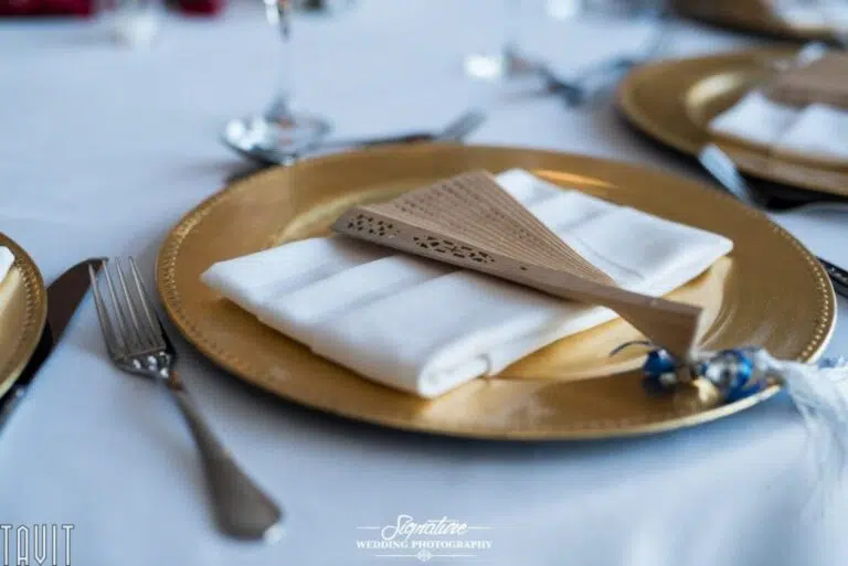 A table setting with a gold charger plate, a folded white napkin, and an intricately designed hand fan on top. Silver cutlery is placed on either side of the plate. The tablecloth is white, and wine glasses are partially visible.