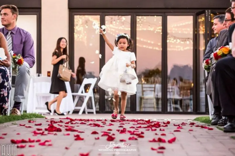 A young girl in a white dress walks down an aisle covered in red rose petals, tossing more petals into the air. Guests in formal attire are seated on either side, and string lights are visible in the background.