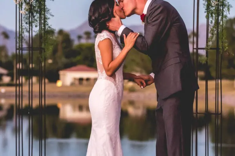 A couple in wedding attire share a kiss under a floral arch by a lakeside, reminiscent of professional headshots. The bride wears a white gown, and the groom is in a gray suit with a red bow tie. The backdrop showcases a calm lake and distant mountains.