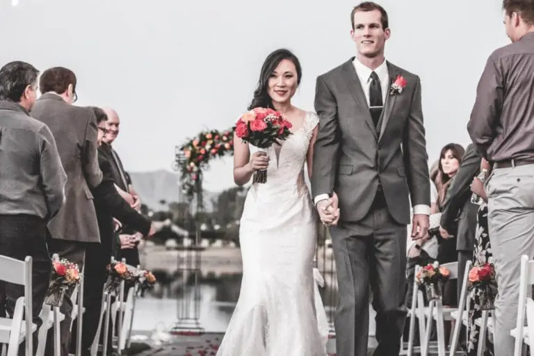 A bride and groom walk hand in hand down an outdoor aisle, decorated with rose petals and surrounded by guests seated on both sides. The groom wears a gray suit, and the bride wears a white gown. They're both smiling. An arch and string lights are in the background, capturing an essence that rivals professional headshots.