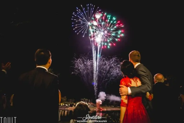 A couple in formal attire enjoys a colorful fireworks display at a corporate event. The woman dazzles in a red dress, while the man has his arm around her, capturing the moment's magic. Others are also seen celebrating in the background, adding to the festive atmosphere.