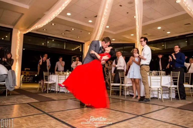 In a large, elegant room adorned with string lights, a couple elegantly dances on the wooden floor. The man in a suit dips the woman in a vibrant red dress, capturing the essence of corporate event photography as onlookers cheer and clap around them at tables with white tablecloths.