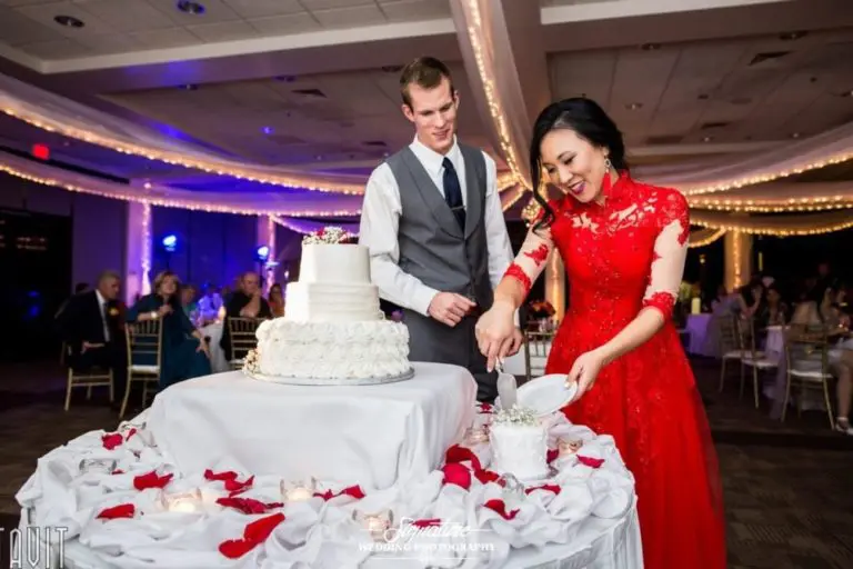 A couple in formal attire cuts a white cake at a wedding reception. The woman dazzles in a red dress, while the man complements her in a gray vest and tie. The elegant table is adorned with red rose petals and white cloth, reminiscent of a beautifully staged corporate event photograph with guests seated in the background.