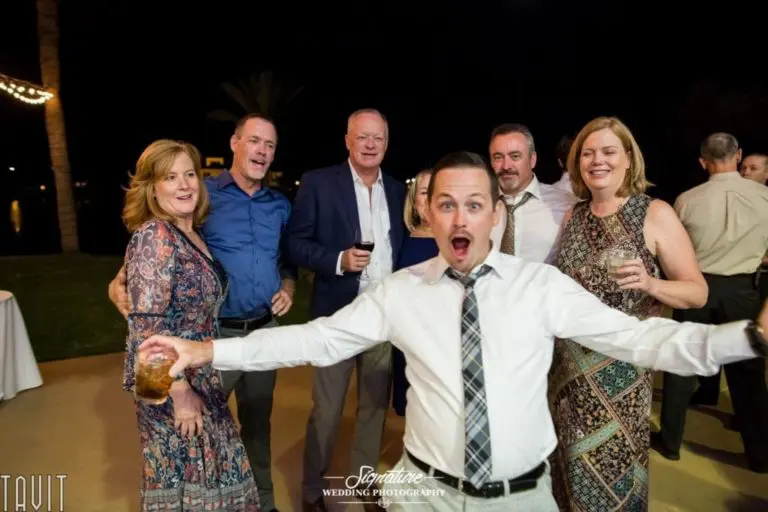 A group of six adults, dressed in formal and semi-formal attire, pose outdoors at night, capturing a moment of corporate event photography. They hold drinks and smile at the camera, while one person in front enthusiastically gestures with arms outstretched.
