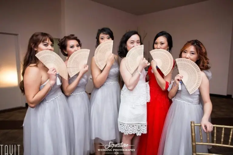 Six women stand together in a room, five wearing light gray dresses and one in a red dress. They are holding fans covering the lower part of their faces, smiling and posing for a corporate event photography session.