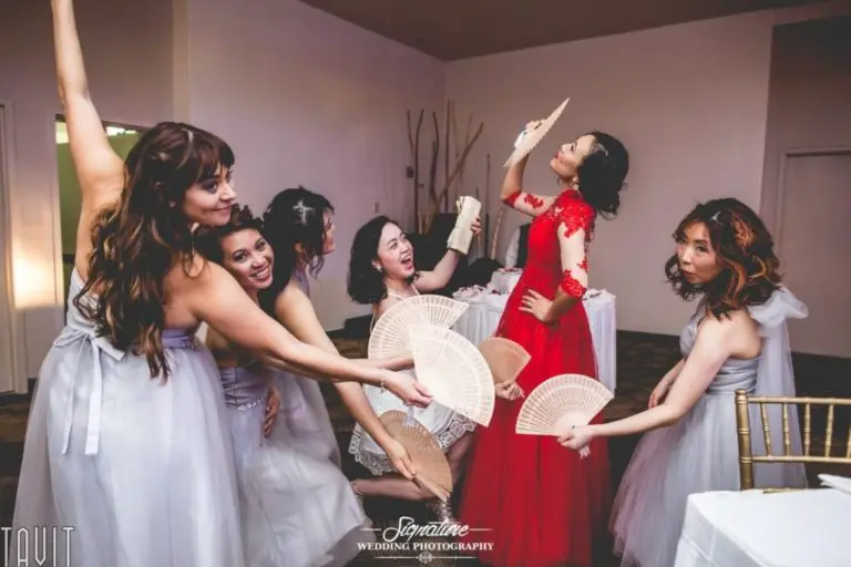 A group of bridesmaids in light gray dresses playfully surround a woman in a red dress, holding fans around her in a fun pose akin to professional headshots. They are indoors, and the atmosphere is lively and joyful, capturing the spirit of corporate event photography.