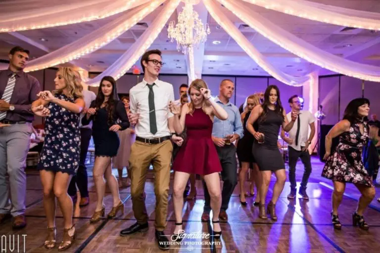 A group of people dance enthusiastically at a wedding reception. Amidst elegant drapery and a chandelier, the scene is reminiscent of a corporate event photography shoot, capturing each joyous moment. Everyone appears to be enjoying themselves on the dance floor in this festive atmosphere.
