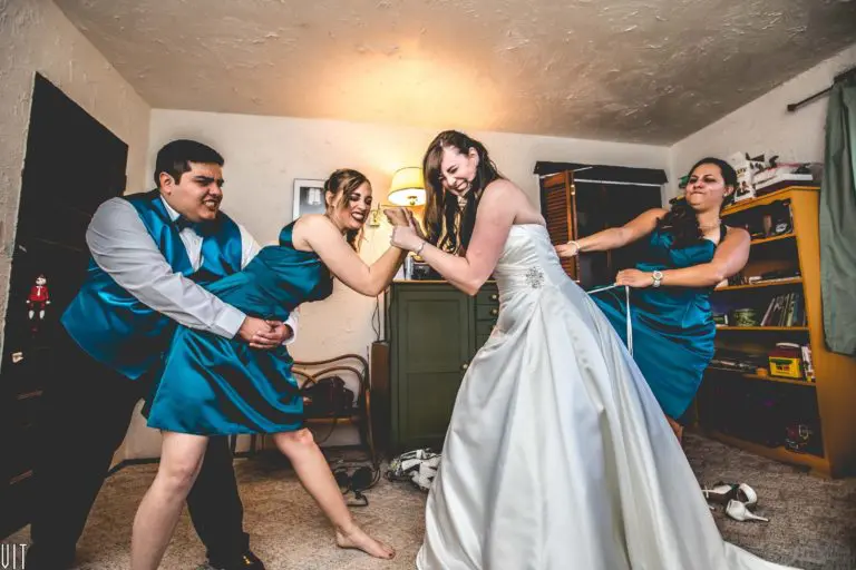 Four people are helping a bride tighten her wedding dress, pulling on the laces and laughing like it’s a fun seminar—not a corporate event—in the living room. Two are in teal bridesmaid dresses, one wears a teal vest and tie.