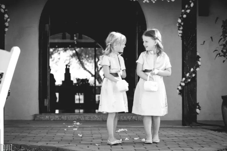 Two young girls in matching dresses and small purses stand facing each other, smiling, outside a decorated building—perhaps after a wedding or seminar. Flower petals are scattered on the ground. The image is in black and white.