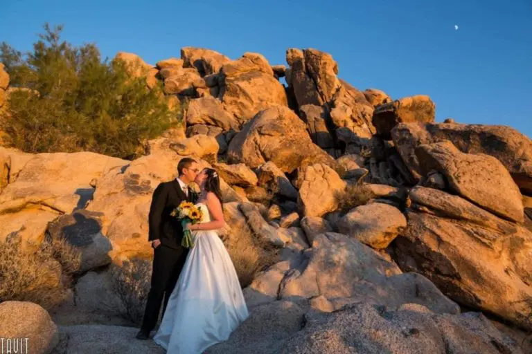 A bride and groom kiss while standing on rocky terrain at sunset, surrounded by large boulders and sparse desert vegetation—turning this landscape into a moment more magical than any seminar or corporate event could offer, beneath a clear sky and small moon.