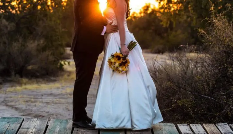 A bride and groom stand close together outdoors at sunset, as if sharing a quiet moment away from a seminar. The bride holds a bouquet of sunflowers, sunlight shining between them and highlighting this romantic scene.