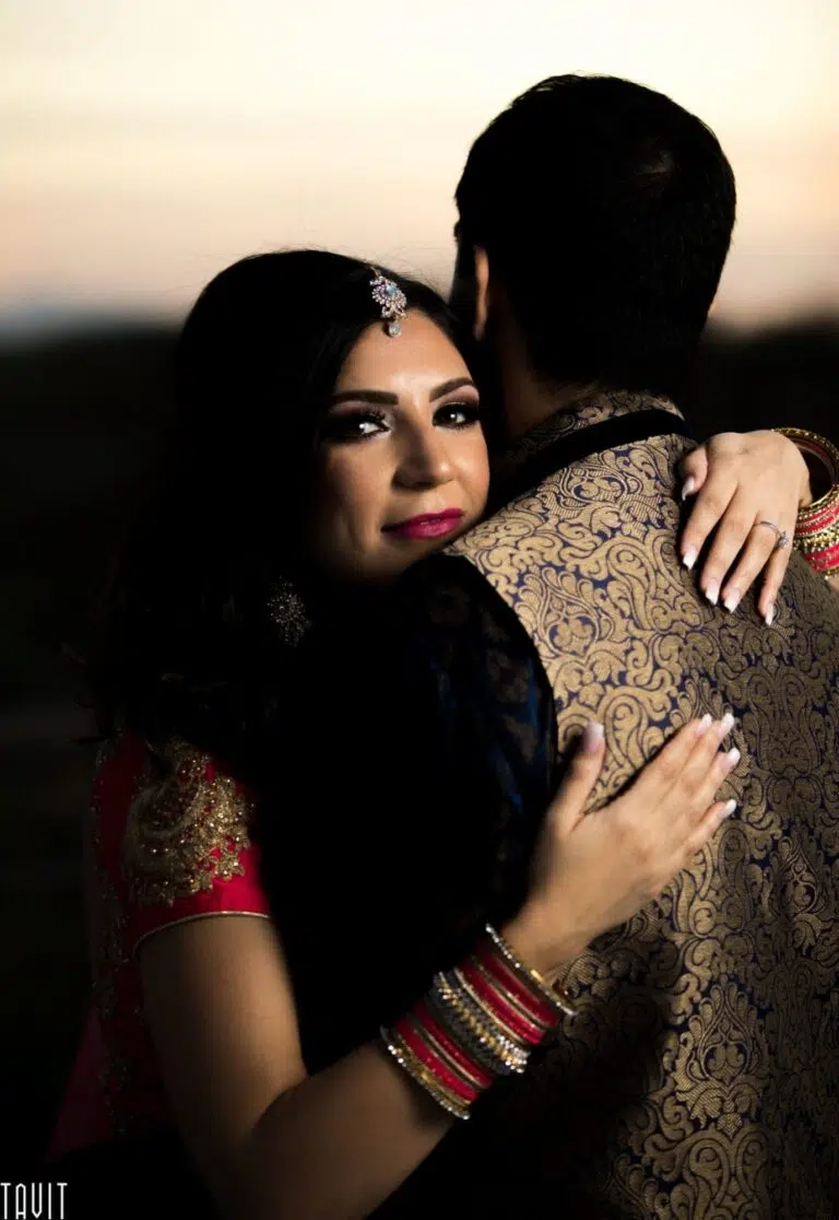 A woman in traditional Indian attire embraces a man from behind, looking at the camera. The man’s back is to the viewer, wearing a gold and blue patterned sherwani—perfect attire for a festive corporate event. The background is softly blurred.