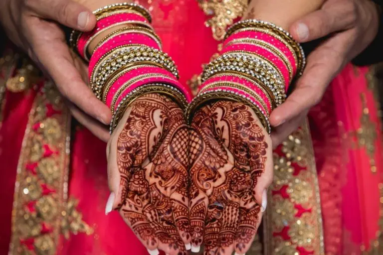 A woman in a red and gold traditional outfit displays intricate henna designs on her palms, with colorful bangles adorning her wrists. A man's hands gently hold hers from behind, capturing a moment of togetherness at a corporate event.