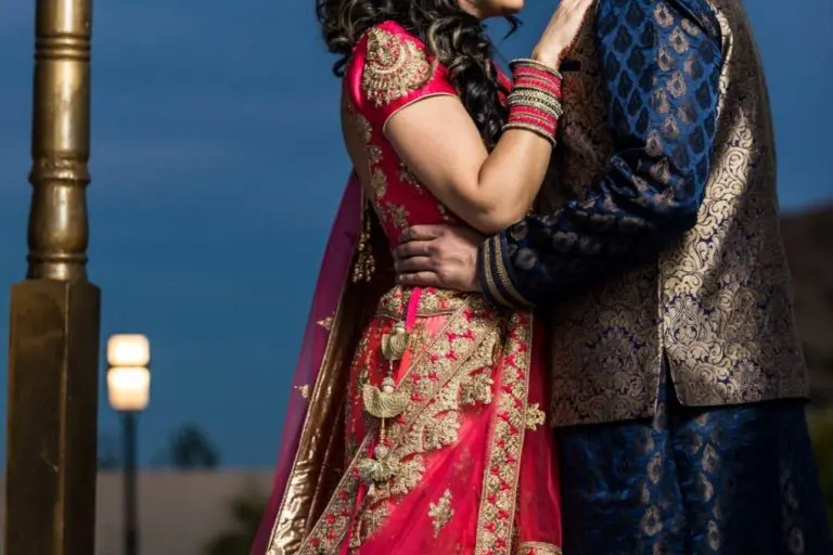 A couple in traditional Indian attire embraces outdoors at dusk, sharing a tender moment after a corporate event; the man kisses the woman's forehead as she smiles with closed eyes, both in intricately embroidered outfits.