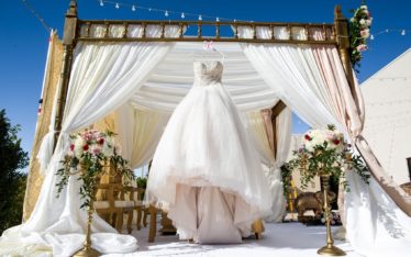 A white wedding dress hangs under a decorated outdoor canopy with flowing white drapes, gold accents, and flower arrangements, creating an elegant scene fit for a celebration or even a stylish corporate event beneath the clear blue sky.