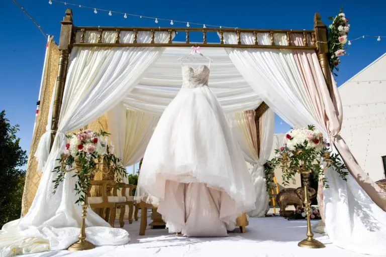 A white wedding dress hangs under a decorated outdoor canopy with flowing white drapes, gold accents, and flower arrangements, creating an elegant scene fit for a celebration or even a stylish corporate event beneath the clear blue sky.