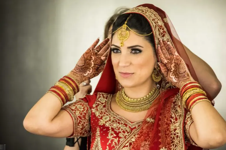 A woman wearing a traditional red and gold bridal outfit with intricate jewelry and henna designs on her hands adjusts her veil, preparing for a cultural appearance at a corporate event.